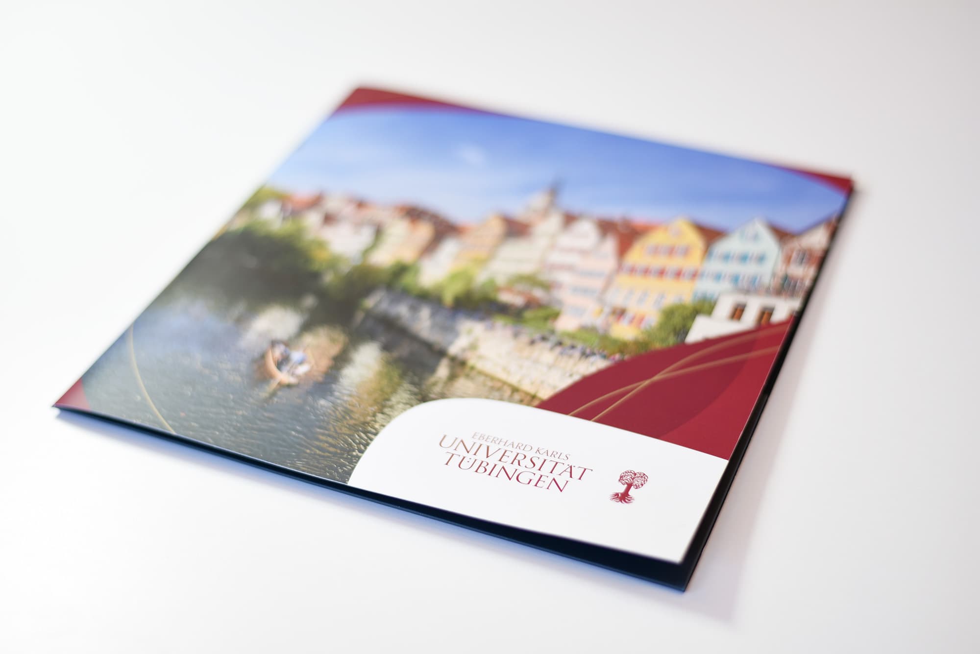 Ein quadratisches Heft mit einem Foto, auf dem ein Fluss und eine Häuserreihe am Fluss zu sehen ist. Vorne rechts eine weiße Box mit dem Text "Eberhard Karls Universität Tübingen" und rechts daneben dem Logo, bestehend aus einem roten Baum.