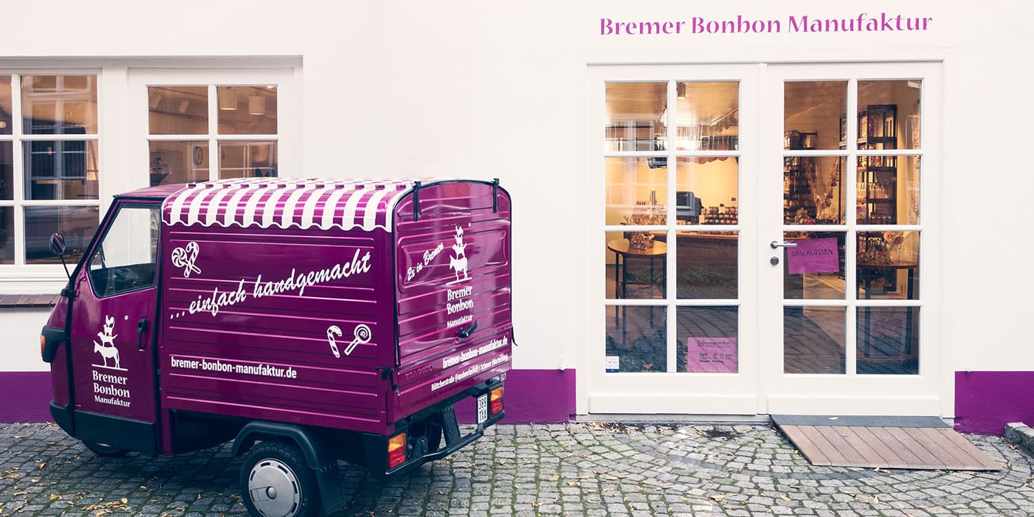 Ein violettes kleines Fahrzeug, eine Ape, das vor einer weißen Hausfassade steht. Die Ape ist von der linken Seite und zum Teil von hinten zu sehen. Auf der Fahrertür ist das Logo der Bremer Bonbon Manufaktur mit den Stadtmusikanten. Auf dem Dach der Transportfläche steht "...einfach handgemacht" und die URL "bremer-bonbon-manufaktur.de". Auf der Rückseite des Fahrzeugs ist wieder das Logo der Bremer Bonbon Manufaktur.