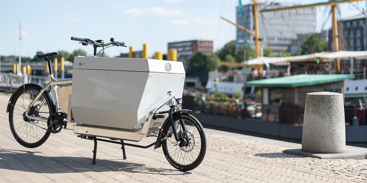 Im Hintergrund das Stadtbild von Bremen an der Weser. Rechts im Hintergrund zwei Bote. Links im Vordergrund ein Lastenrad mit silbernem Rahmen, schwarzen Reifen und einer weißen großen Box vorne zwischen Lenker und Vorderreifen.