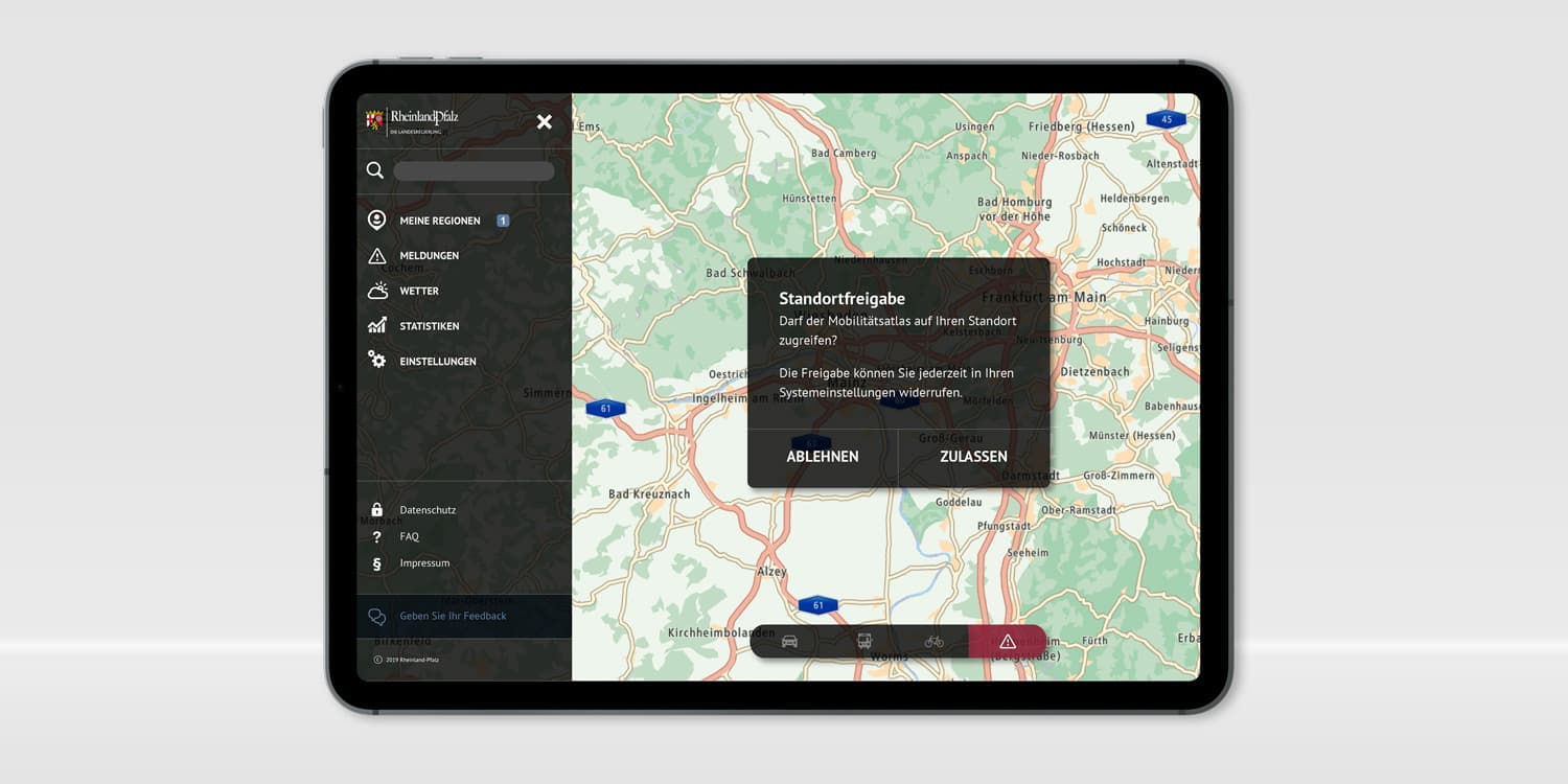 Ein Tablet, auf dem der Mobilitätsatlas geöffnet ist. Im linken Drittel befindet sich das schwarze Menü. Oben links das Logo von Rheinland-Pfalz, darunter eine Suchleiste und darunter die Menüpunkte "Meine Regionen", "Meldungen", "Wetter", "Statistiken", "Einstellungen", "Datenschutz", "FAQ", "Impressum" und ein Button "Geben Sie Ihr Feedback". Auf dem Rest des Bildschirms ist eine Landkarte zu sehen mit der Region Franfurt am Main. Darüber ist ein schwarzes Popup geöffnet mit dem Text "Standortfreigabe: Darf der Mobilitätsatlas auf Ihren Standort zugreifen? Die Freigabe können Sie jederzeit in Ihren Systemeinstellungen wiederrufen." und den beiden Schaltflächen "Ablehnen" sowie "Zulassen".