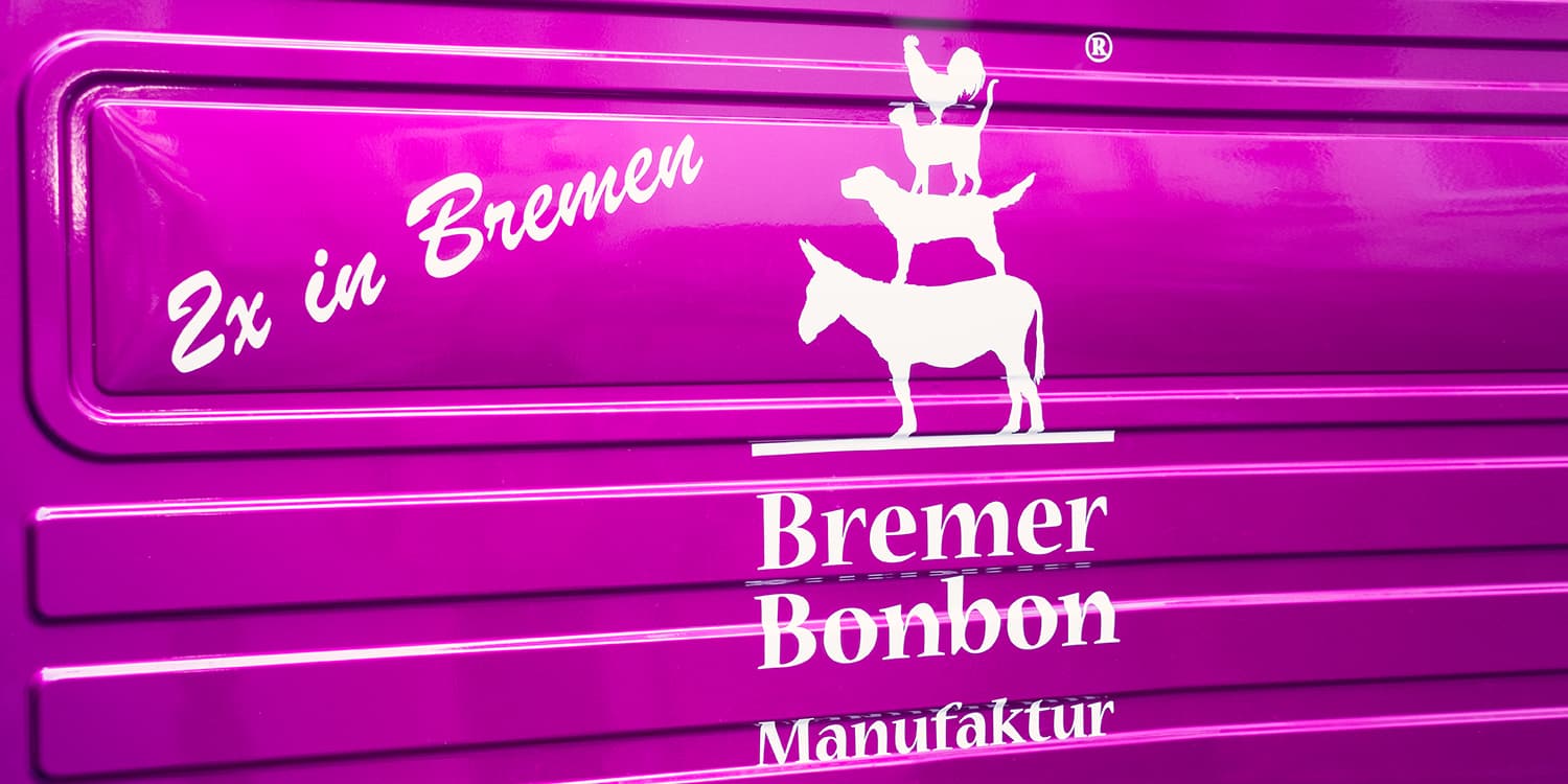 Ein violetter metallischer Untergrund. Oben links steht in weiß "2x in Bremen", in der Mitte befindet sich das weiße Logo der Bremer Bonbon Manufaktur mit den Bremer Stadtmusikanten.